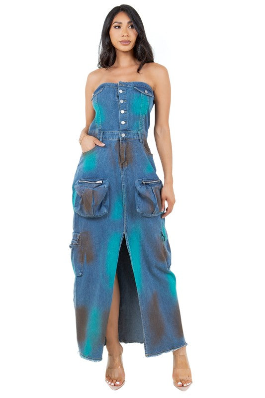 Model wearing a strapless, button-front denim maxi dress with a front slit. The dress features large utility-style pockets on both sides and a distressed, tie-dye pattern in teal and brown shades. Paired with clear high-heeled sandals