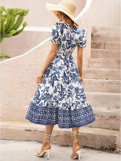 Woman in a blue and white floral dress with a V-neck, short sleeves, and a flowy skirt, wearing a straw hat, strappy heels, and statement earrings, posing on concrete steps.