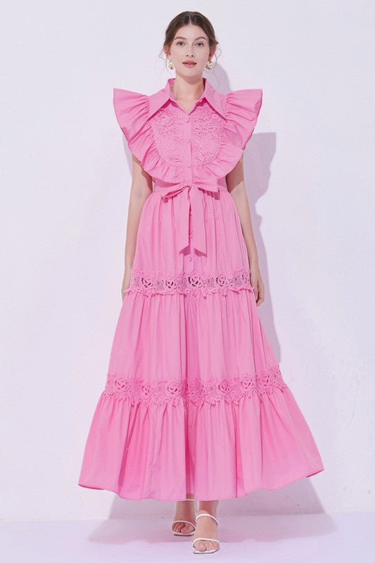 Woman wearing a sleeveless embroidered maxi dress with dramatic ruffle sleeves, tiered lace skirt, collared neckline, and waist tie, styled with heels.

