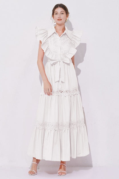 Woman wearing a sleeveless embroidered maxi dress with dramatic ruffle sleeves, tiered lace skirt, collared neckline, and waist tie, styled with heels.