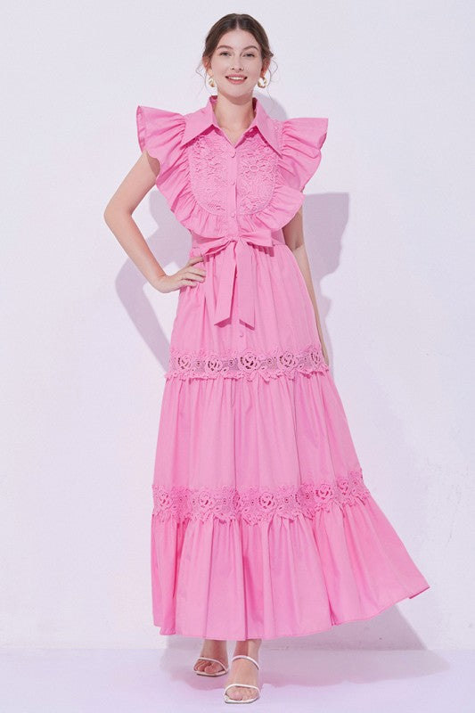 Woman wearing a sleeveless embroidered maxi dress with dramatic ruffle sleeves, tiered lace skirt, collared neckline, and waist tie, styled with heels.
