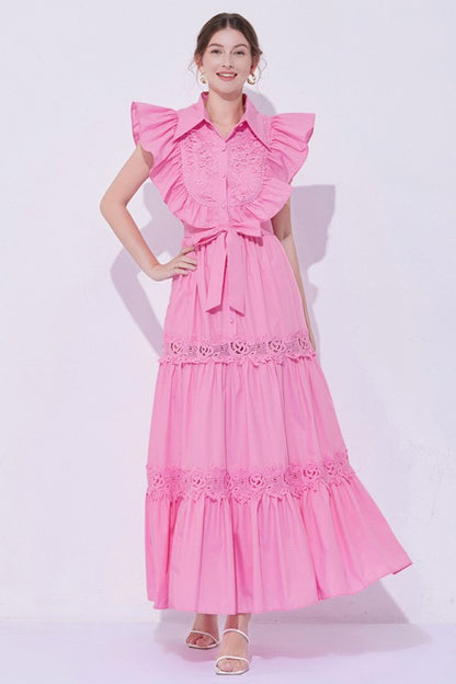 Woman wearing a sleeveless embroidered maxi dress with dramatic ruffle sleeves, tiered lace skirt, collared neckline, and waist tie, styled with heels.