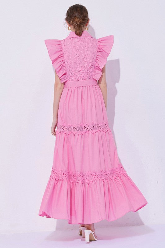 Woman wearing a sleeveless embroidered maxi dress with dramatic ruffle sleeves, tiered lace skirt, collared neckline, and waist tie, styled with heels.