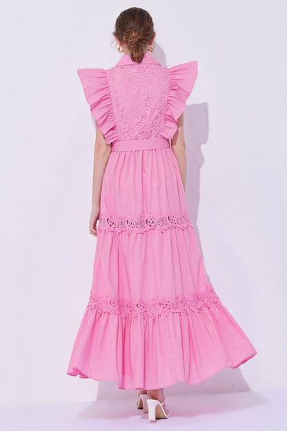 Woman wearing a sleeveless embroidered maxi dress with dramatic ruffle sleeves, tiered lace skirt, collared neckline, and waist tie, styled with heels.