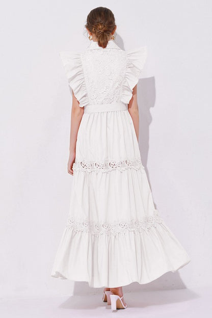 Woman wearing a sleeveless embroidered maxi dress with dramatic ruffle sleeves, tiered lace skirt, collared neckline, and waist tie, styled with heels.