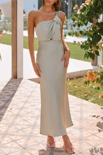 A light taupe, satin-like, sleeveless maxi dress with a draped, twisted front detail and a sleek, form-fitting silhouette.