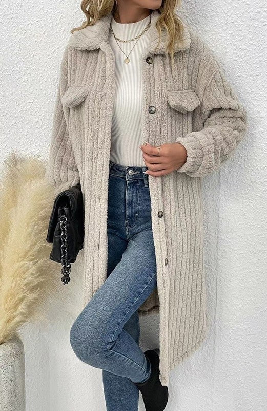 Person wearing a beige ribbed knit cardigan over a white sweater and blue jeans, standing against a white wall.