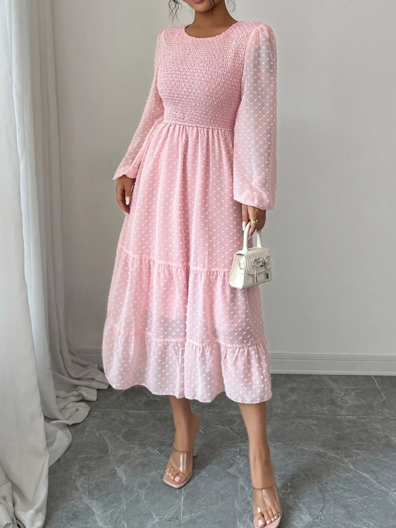 A pink dress features a fitted bodice with long sheer sleeves and a tiered flowing skirt that falls tomid-calf.