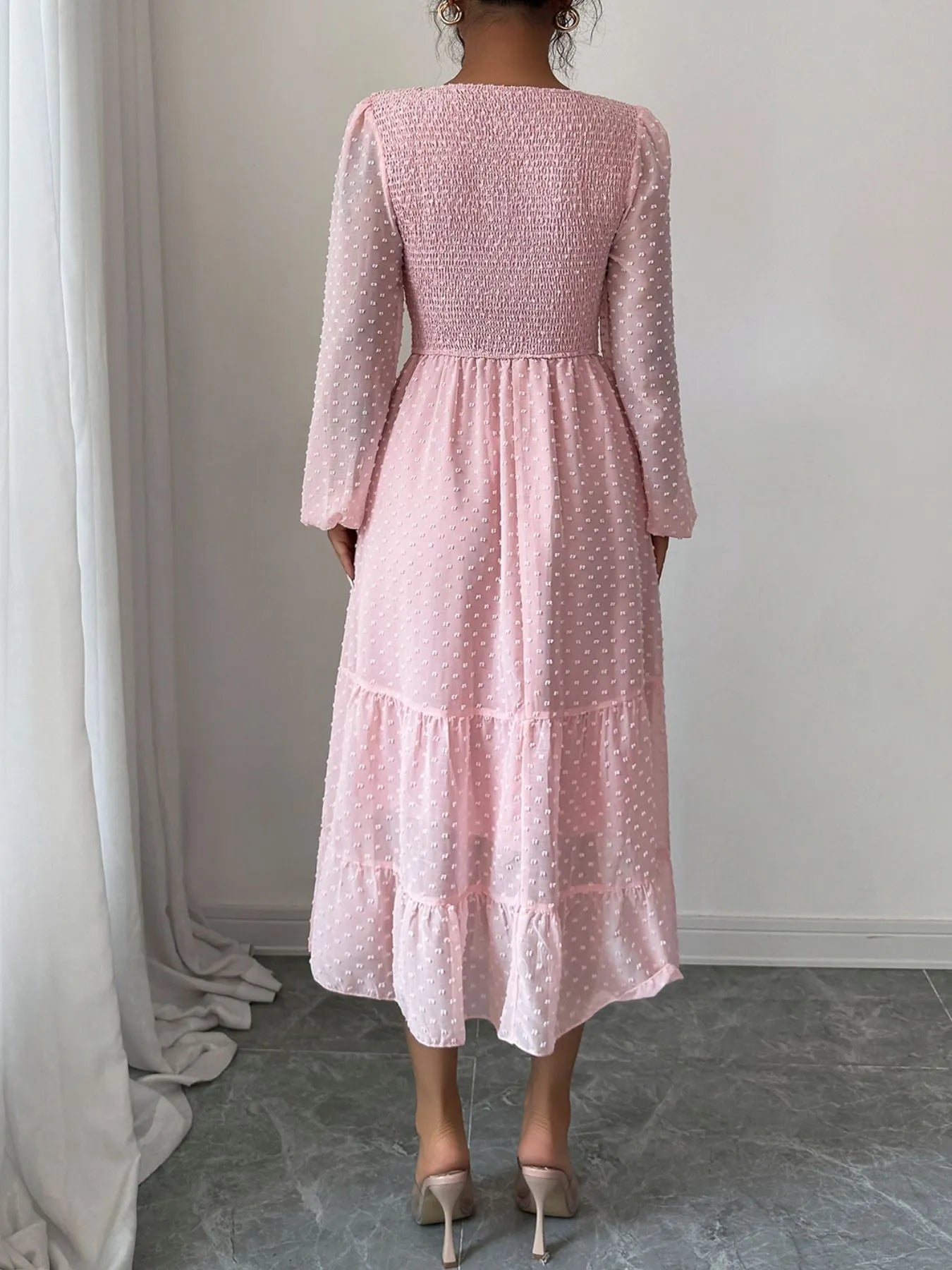 A pink dress features a fitted bodice with long sheer sleeves and a tiered flowing skirt that falls tomid-calf.