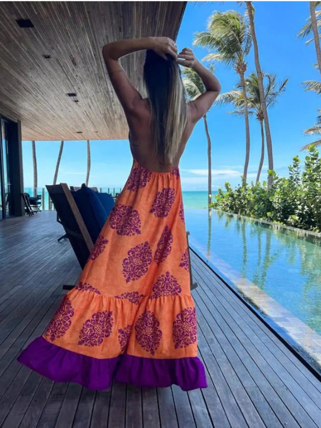 Woman wearing a floral halter-neck maxi dress with deep V-neckline and solid trim, standing by a tropical infinity pool
