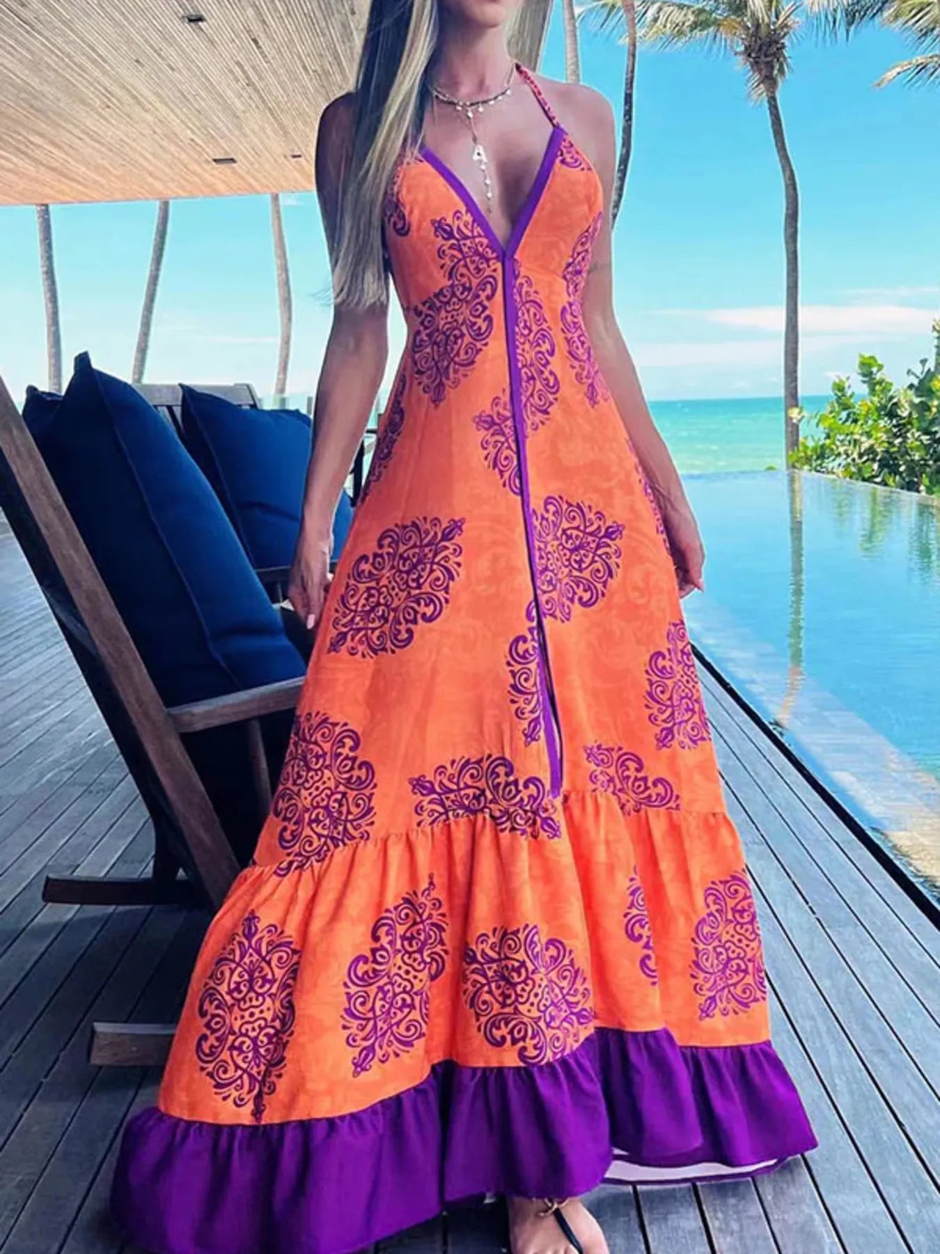 Woman wearing a floral halter-neck maxi dress with deep V-neckline and solid trim, standing by a tropical infinity pool