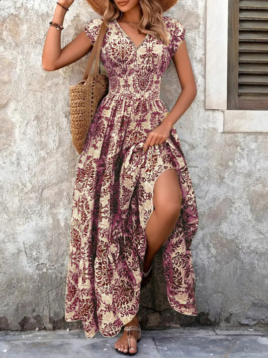 Woman wearing a maroon floral V-neck maxi dress with cap sleeves and high slit, styled with a straw hat and handbag in a tropical outdoor setting.