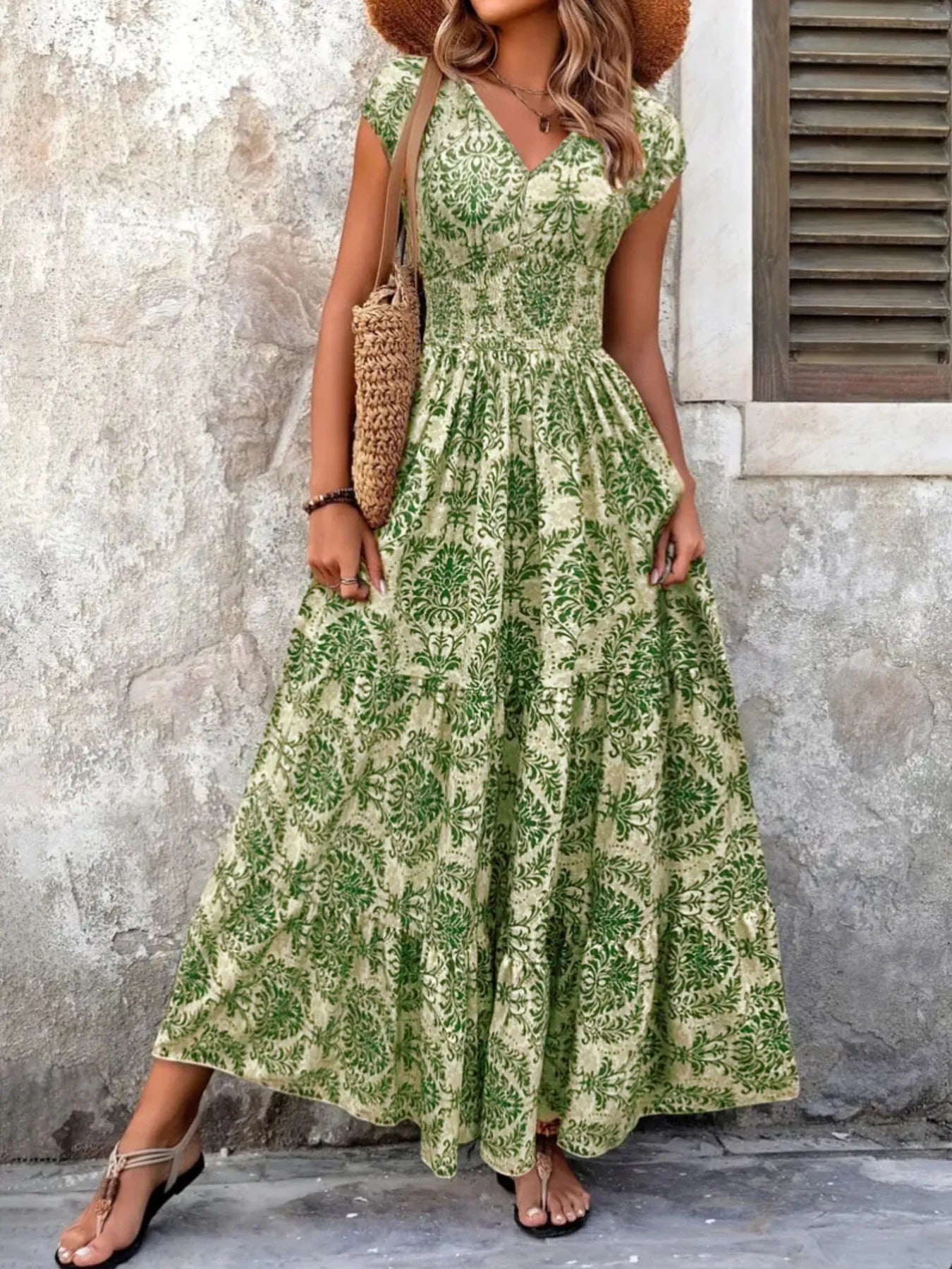 Woman wearing a maroon floral V-neck maxi dress with cap sleeves and high slit, styled with a straw hat and handbag in a tropical outdoor setting.