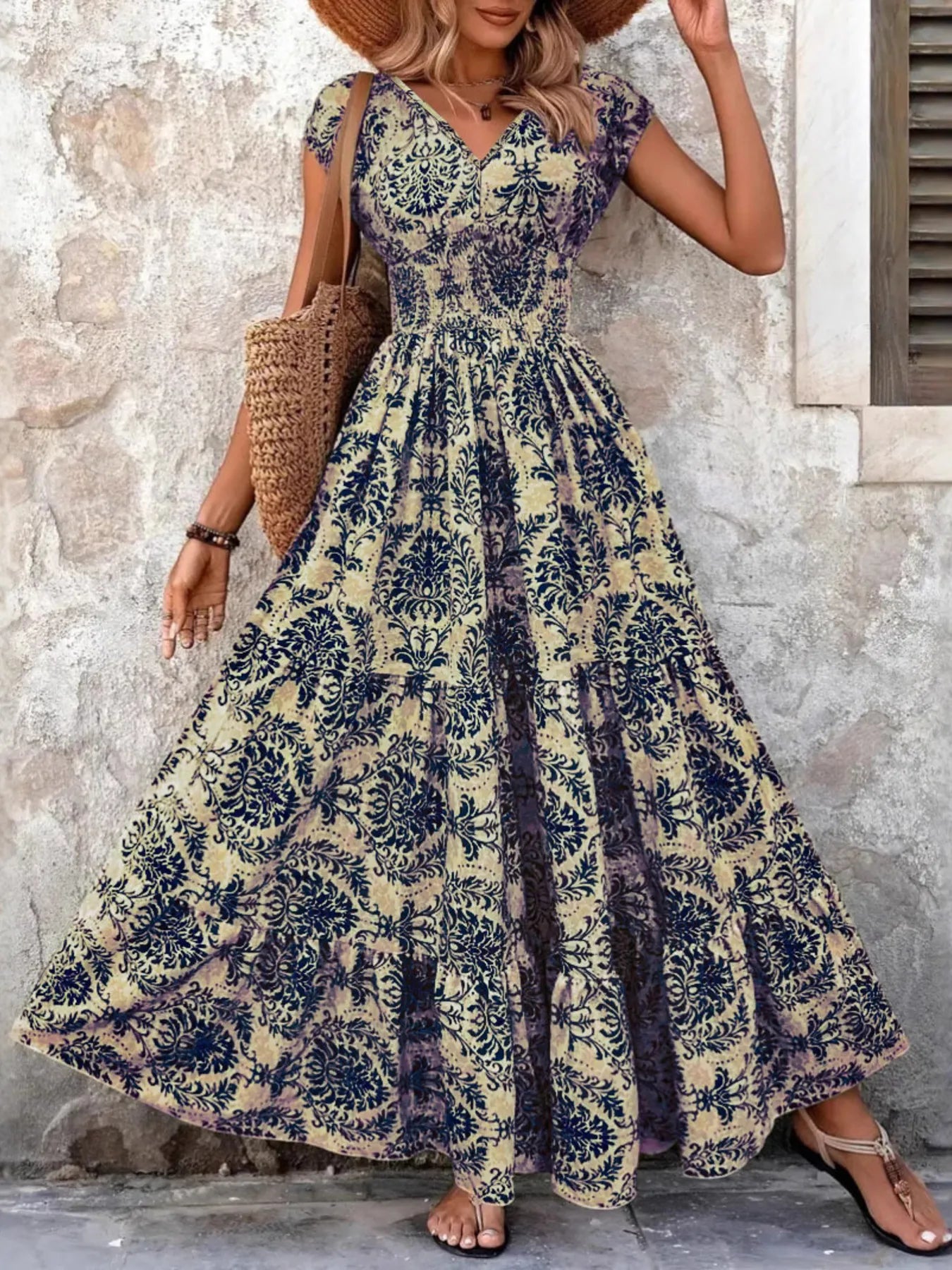 Woman wearing a maroon floral V-neck maxi dress with cap sleeves and high slit, styled with a straw hat and handbag in a tropical outdoor setting.