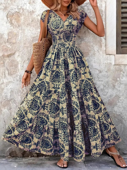 Woman wearing a maroon floral V-neck maxi dress with cap sleeves and high slit, styled with a straw hat and handbag in a tropical outdoor setting.