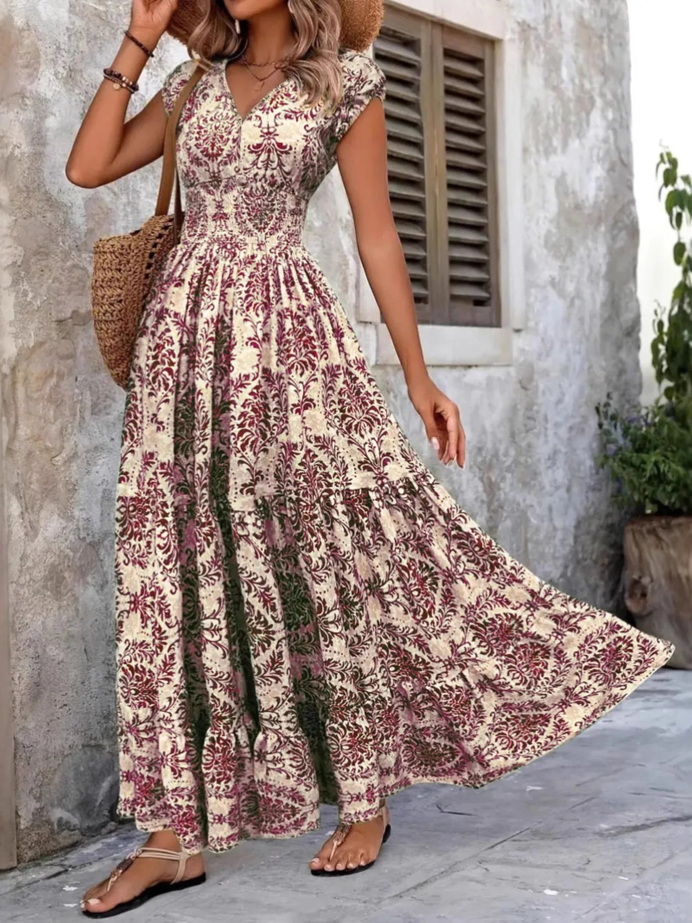 Woman wearing a maroon floral V-neck maxi dress with cap sleeves and high slit, styled with a straw hat and handbag in a tropical outdoor setting.
