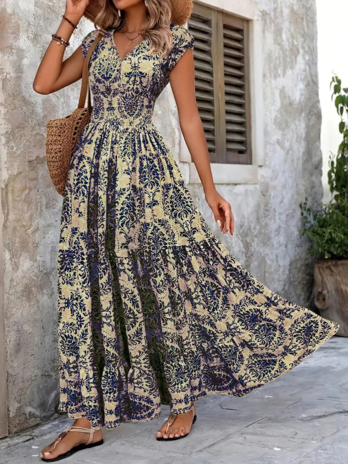 Woman wearing a maroon floral V-neck maxi dress with cap sleeves and high slit, styled with a straw hat and handbag in a tropical outdoor setting.