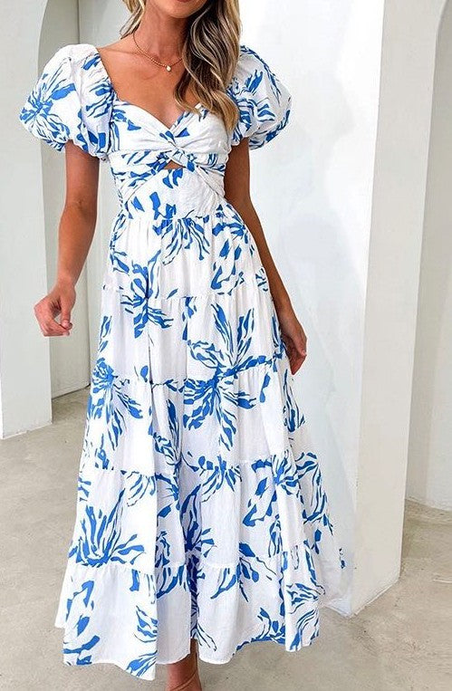 A white maxi dress with a blue botanical print features short puff sleeves and a wrap-style bodice that ties at the waist.