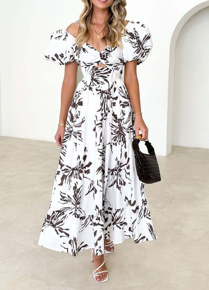 A white maxi dress with a blue botanical print features short puff sleeves and a wrap-style bodice that ties at the waist.