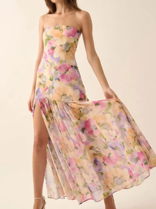 Woman wearing a pastel floral strapless dress with a high side slit, holding out the skirt to showcase its flowy design against a plain white background.
