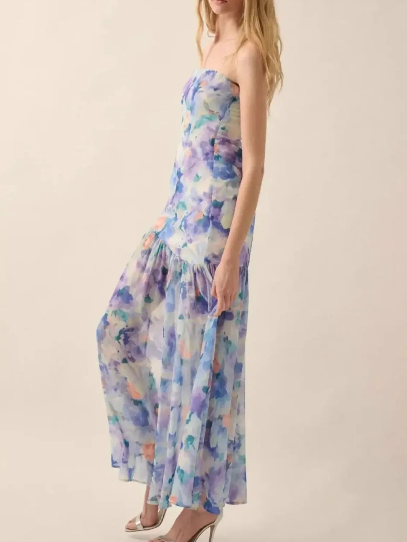 Woman wearing a pastel floral strapless dress with a high side slit, holding out the skirt to showcase its flowy design against a plain white background.
