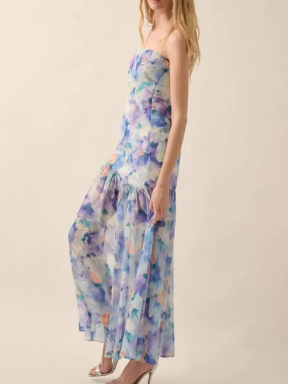 Woman wearing a pastel floral strapless dress with a high side slit, holding out the skirt to showcase its flowy design against a plain white background.
