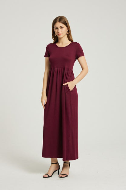 A woman in a maroon dress sits on a wooden chair with a black backrest, posing against a plain background.